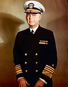 Man in dark blue suit and tie, wearing peaked cap and two rows of ribbons