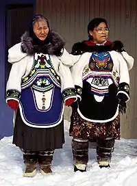 Two Inuit women in clothing covered with detailed beadwork and colourful fabric designs. They have fur ruffs on the hoods of their parkas, cloth skirts beneath their parkas, and fur boots.