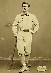 A baseball player is standing, facing the camera, with the top of a baseball bat in his right hand, and the bottom resting on the ground