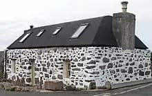 A small house with a black roof and a small chimney. The walls are constructed of stone and mortar; the former are undressed whilst the latter are painted white, creating a spotted effect.