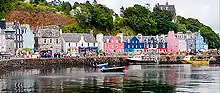  A row of brightly coloured stone buildings front a harbour with a tree-clad cliff behind. The houses are painted predominantly in shades of pink, blue and white. In the foreground, small boats lie in the water by the shelter of a stone pier.