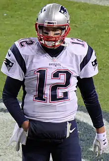 American football quarterback, Tom Brady, walks onto a football field while holding a gray helmet. He is wearing a white New England Patriots jersey with a blue number "12" and blue pants.