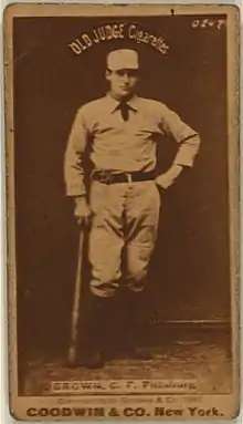 A baseball player is standing, holding a baseball bat down his right side.