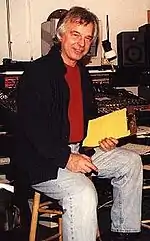 A gray-haired man with a red shirt and jacket sitting on a chair