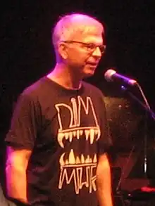 A gray-haired man with glasses and a black shirt standing in front of a microphone