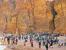 A large variety of people taking photographs of something just beyond the camera, in a canyon with a rocky rear wall