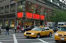 A street view of a store front prominently features a yellow taxi in front of the store. The walls contain clear windows with the phrase "Tower Records – Video" in bright orange lights.