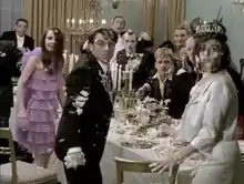 A group of finely dressed dinner guests are gathered around a messy dining table. They are covered with globs of food. All their heads are turned toward the viewer with shocked expressions.