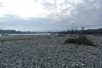 Photo shows a river bordered by an immense bed of white stones. There are mountains in the distance.