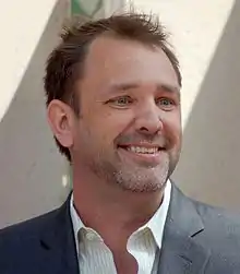 A brown-haired man wearing a white shirt and gray jacket smiles.