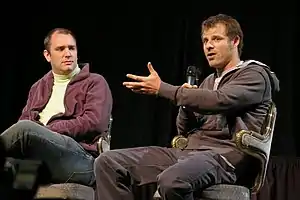 Two men sit in chairs. The man on the right holds a microphone and gestures with his left hand.