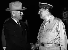 Truman in a dark suit and tie and light hat shakes hands with MacArthur, in uniform wearing a shirt but no tie and his rumpled peaked cap.