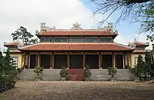 A temple with red tiles, pillars holding up the two levels. There are concrete steps leading to the temple, and potted shrubs in front of them. A stone courtyard is in the foreground, flanked by more shrubs.