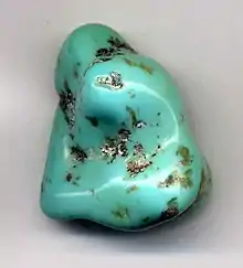 A polished, freeform cabochon of turquoise blue with brown dots of matrix inclusions.