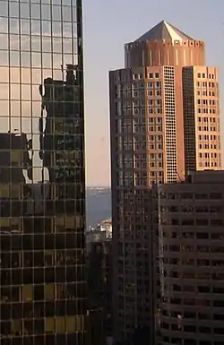 Aerial view of a round building with a light-red facade, topped with a pyramid-like cap; in the foreground is a shorter building with an all-glass facade