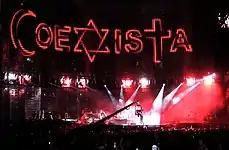 A view of the stage being filmed, with "COEXISTA" spelled out on the video screen, using an Islmaic crescent as a "C", a Star of David as an "X", and a Christian cross at a "T". A camera in the middle shows the concert being filmed.