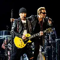 The Edge and Bono clothed in leather jackets, as the Edge holds a guitar vertically. A large dangling light bulb hangs between them.