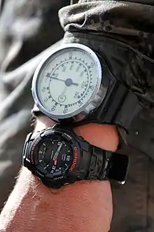 A diver's left wrist, wearing a diving watch and mechanical depth gauge with a needle indicator