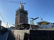 A high-rise building under construction with tall crane along side a light rail station platform