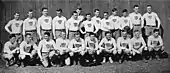 A group of 23 young men arranged in two rows—thirteen standing in back and ten squatting in front—in rugby uniform: white sweaters emblazoned with vertically striped shields, black belts, and light colored shorts.