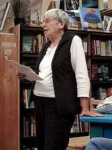 Photograph of Ursula K. Le Guin standing and reading aloud in a bookshop