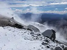 A snow-covered mountain slope is surmounted by steam clouds; mountainous landscape in the background