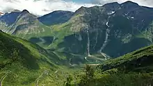 A small road climbs a mountainside in S-bends surrounded by green mountainsides and barren peaks.