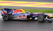 A figure, wearing a black helmet design, is driving a Formula One car that is of a blue colour design, with red, light blue and white stripes adorned on the car's front section.