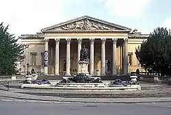  A Palladian style nineteenth century stone building with a large colonnaded porch. In front a large metal statue on a pedestal and fountains with decorations.