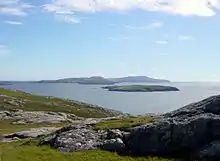 A rocky moorland in the foreground and low, dark islands against a blue sky in the background.