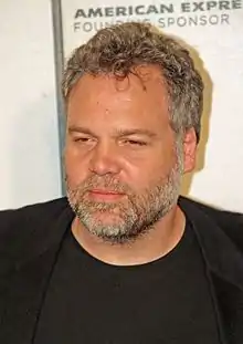 A man wearing a black shirt, donning a gay beard and pepper-colored hair, stands in front of a promotional image for a credit card company.