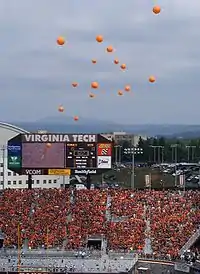 Orange balloons rising above Lane Stadium, with everyone in the stands wearing maroon or orange, and the stadium scoreboard in the background.