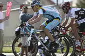 Two cyclists, one wearing a blue and white jersey with yellow trim and the other, to his right, a black and white jersey with red trim, race down a road while spectators watch from beside them