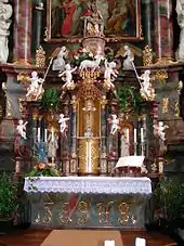 The altar and the reredos that rises behind it together are an example of German Baroque church fitting. They have polychrome marbled surfaces of pink and grey which match the columns of the church. The reredos is heavily architectonic with columns, niches, scrolls and pediments of different shapes which rise in stages to a plinth on which rests a Medieval statue of the Virgin Mary grieving over the body of Christ. On the level below this is a statue of the Lamb of God. In the niche beneath is a silver cross of Medieval design. Beneath this is a gilt metal sacrament cupboard. The central objects are surrounded by a profusion of shiny white stucco cherubs, angels and saints. Across the front of the marble altar is the name "Jesus" is elaborate capital letters. The altar is decked with a lace-edged cloth, flowers and a Bible.