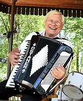 Seated, smiling man with wire-frame glasses, playing a piano accordion that is decorated with the name "OSTANEK".