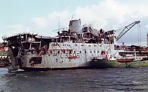 A warhead with the superstructure collapsing and largely gutted by fire. A much smaller green-hulled vessel is moored alongside.