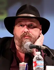 A man in a beard and wide-brimmed hat addresses a crowd at a convention.