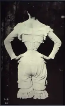 A view of a woman in a corset and bloomers from the back with her hands on her hips. She has a very small waist.