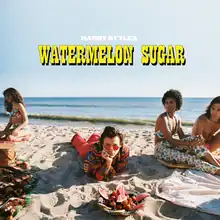 Harry lies prone on the beach sand with a fruit bowl in front, females to the left and right, and the ocean behind.