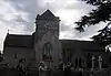 Gray stone building with arched windows. A central tower has a clock on the near face and is surmounted by a weather vane.