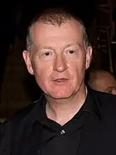 Headshot of a man wearing an open-necked black shirt, not smiling or looking directly at camera