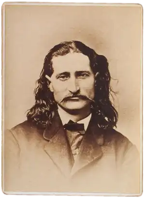 A slightly smiling man dressed in an overcoat and sporting a mustache and shoulder-length, curly hair stares ahead.