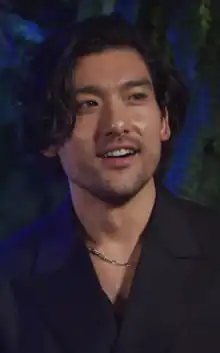 Will Sharpe wearing a dark shirt and a chain on his neck, grinning and looking left of camera