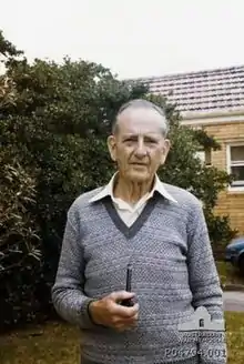 Informal portrait of an older man. He is standing and is only visible from the waist up. His left arm is straight down, while the right is holding a smoking pipe near his chest. He has grey hair, and is wearing a blue-grey sweater with a white collared shirt underneath. A bushy, green tree and part of a house is seen behind him.