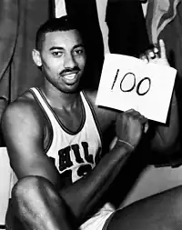 Wilt Chamberlain, an African American man, is shown sitting down in his Philadelphia Warriors jersey while holding up a piece of paper with the number 100 written on it. The photograph was taken directly after the game and is in black and white.