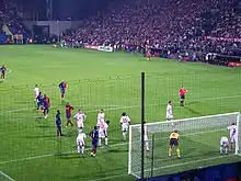 A photograph from behind the goal shows Barcelona and Wisła Kraków players gathering in the near penalty area for a corner kick. Wisła Kraków players are standing closer to the goal, and the referee is standing on the edge of the penalty area.