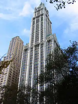 Color photo of the Woolworth Building, a skyscraper, with trees in the foreground and a shorter building to the left