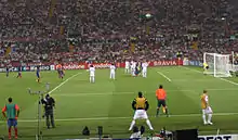 A man in a blue-and-red shirt kicks a football past a row of men in white shirts towards a goal.