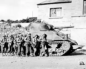 A tank is parked in front of a house. Seven soldiers stand in front of the tank.