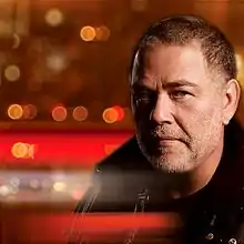 close-up of Yehuda Poliker wearing a black jacket, peering intently at camera, with blurred city lights in background